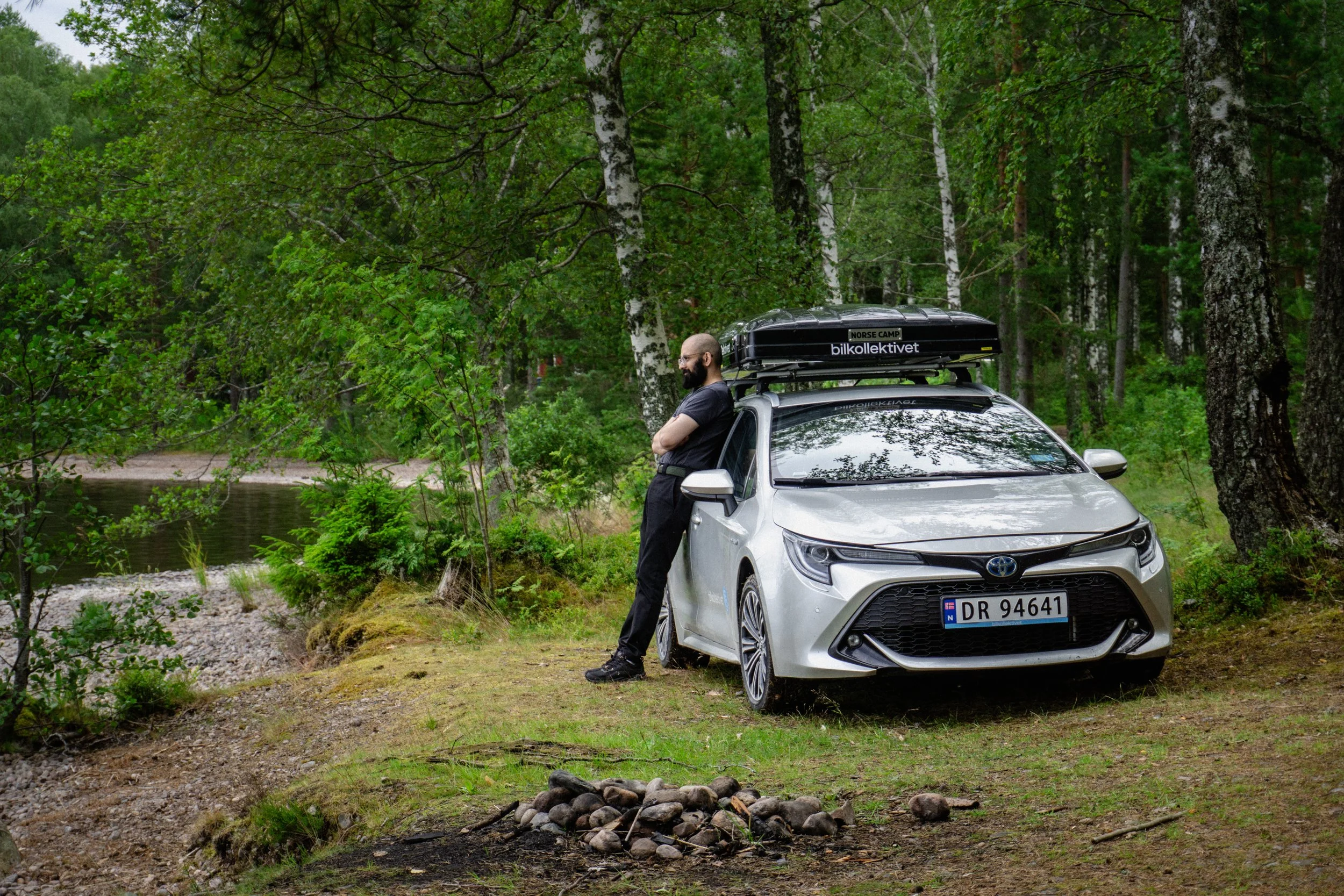 En mann står med armene korslagt ved siden av en hvit Toyota Corolla med en takboks på toppen, i en skog ved en liten bekk, omgitt av grønne trær og vegetasjon.