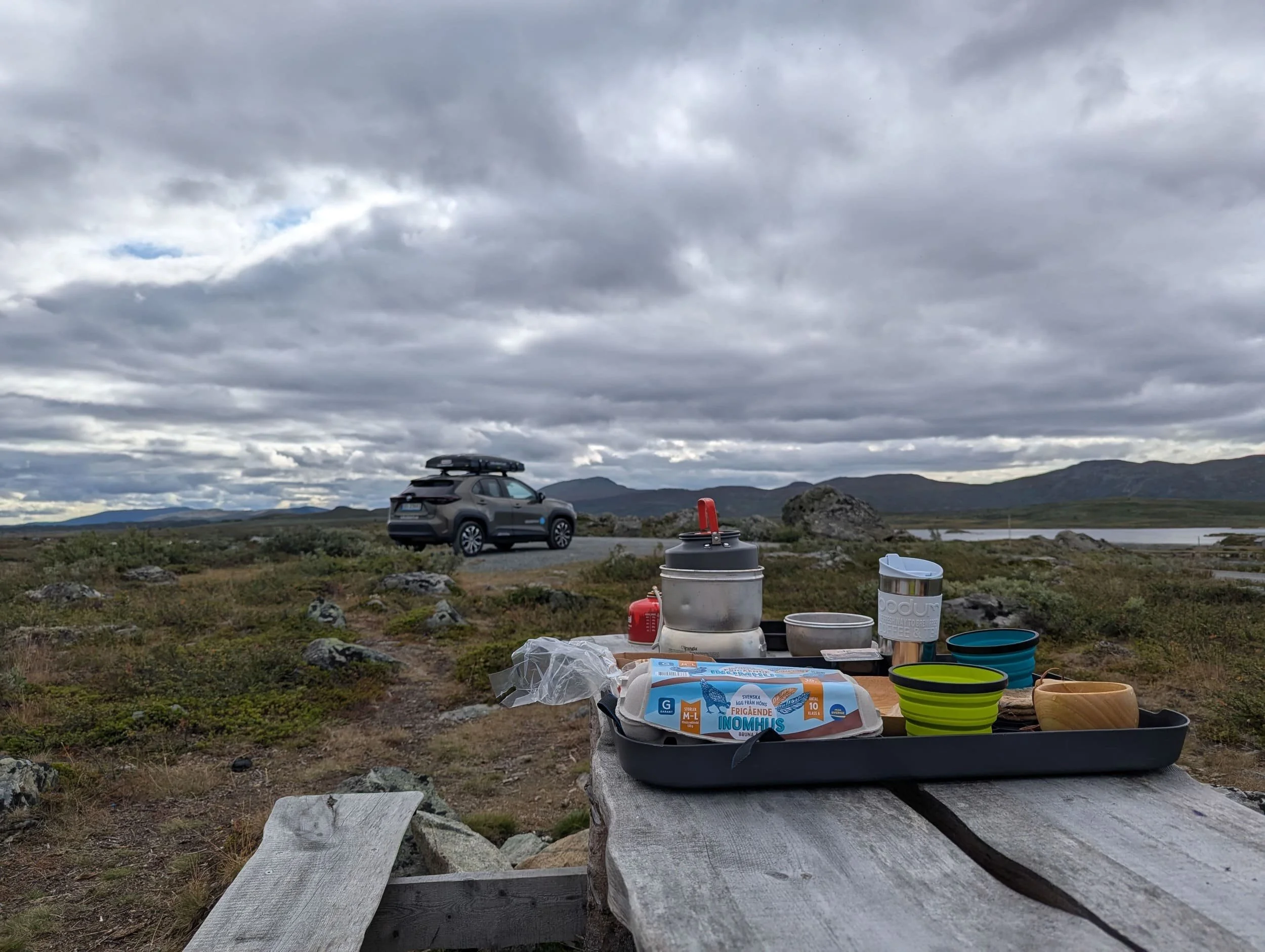 Utsikt over en parkeringsplass med en bilkollektivet bil med taktelt og campingutstyr, mot en overskyet himmel og fjell i bakgrunnen, med en utendørs bord med mat og kopper i forgrunnen.