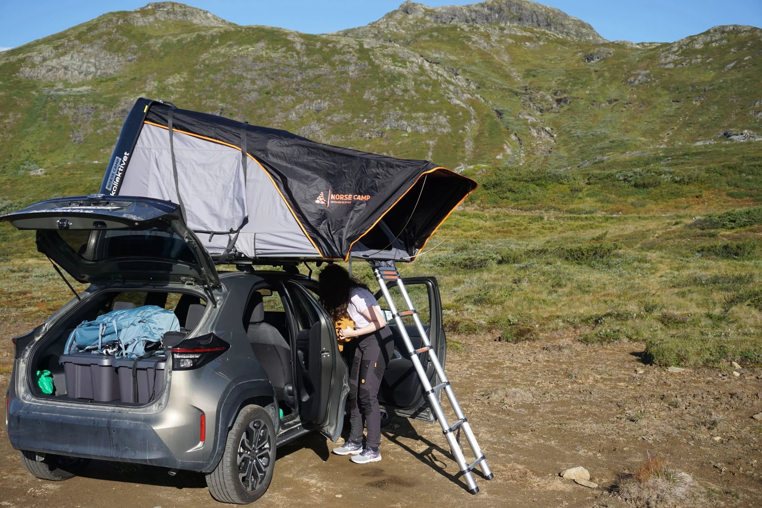 To personer pakker utstyr fra en bil med campingutstyr i en fjellandskap med grønne åser og kalde fjell. En person står på bakken ved siden av bilen, mens en annen klatrer opp en sammenleggbar trapp. Det er et telt festet på taket av bilen.