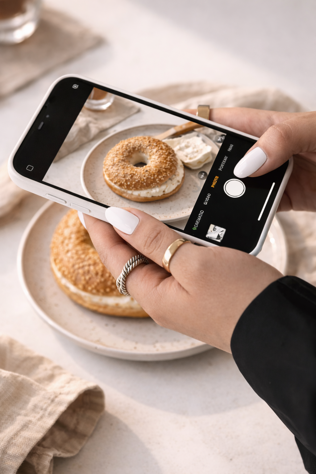 Close-up of hands with white manicured nails holding a smartphone and taking a photo of a sesame bagel with cream cheese on a plate.