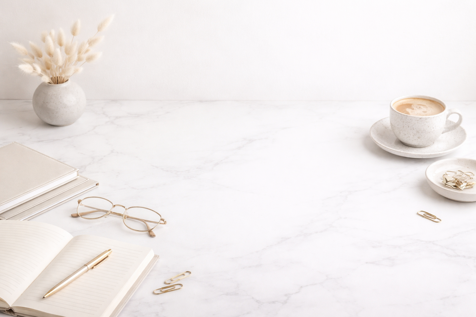 Minimalist neutral workspace with a white marble desk, open notebook and gold pen in the foreground, beige notebooks and gold-rimmed glasses to the left, a small ceramic vase with dried bunny tails, and a speckled coffee cup on a saucer to the right,