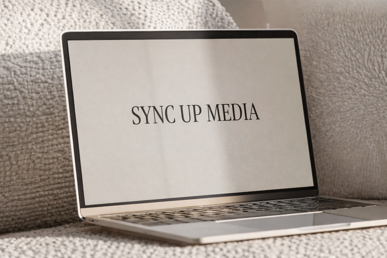 Open laptop resting on a textured beige sofa displaying “SYNC UP MEDIA” on the screen.