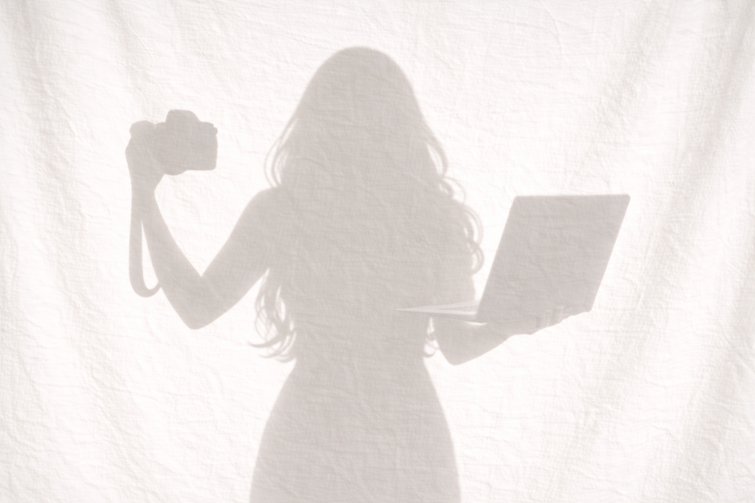 Shadow silhouette of a woman holding a laptop and a camera cast onto a bright white linen background with visible fabric texture.