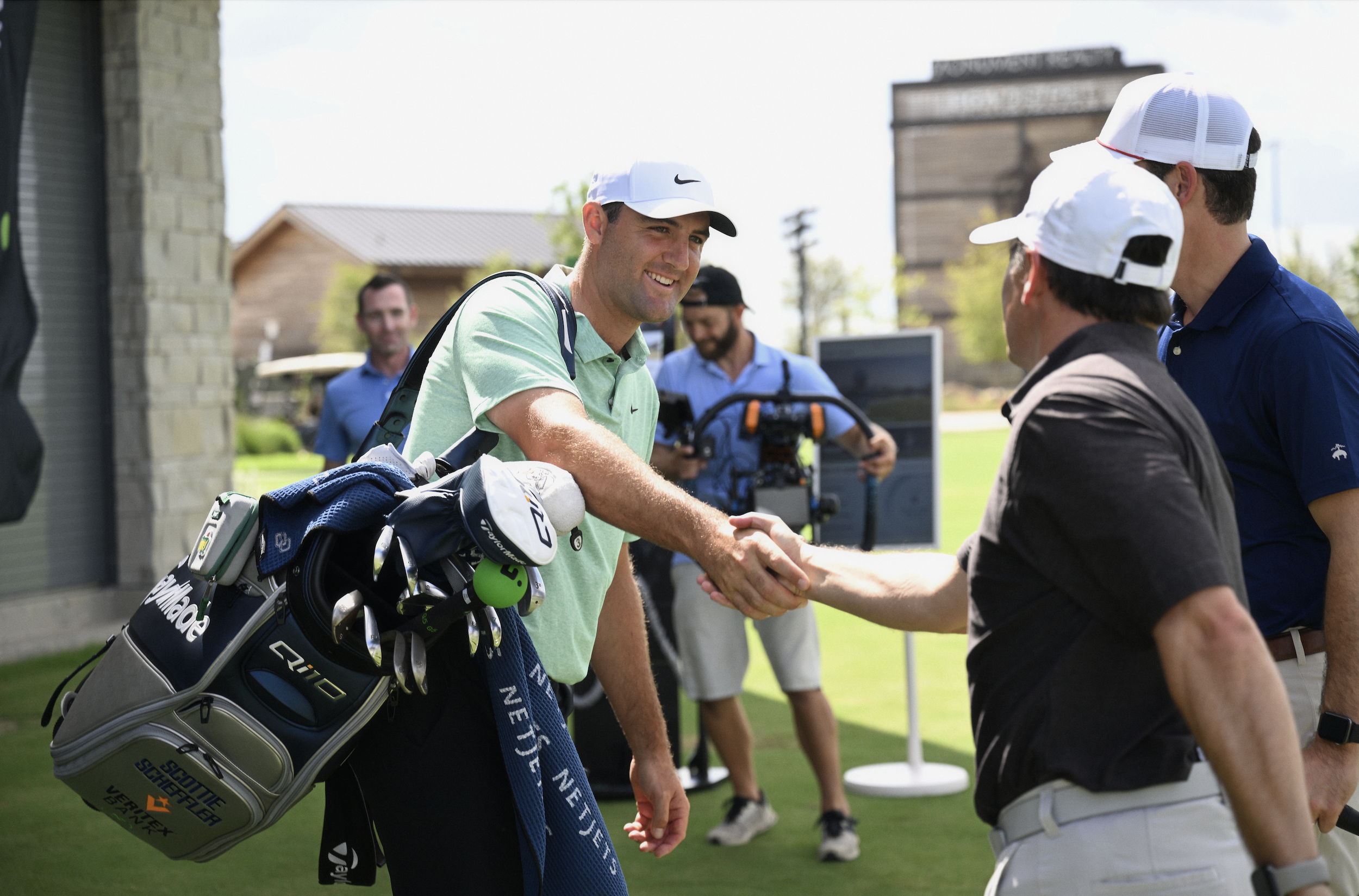 A man in a green shirt and white cap shakes hands with another man wearing a dark shirt and white cap on a golf course. A golf bag with equipment is visible, and several people are in the background, including one holding a video camera.