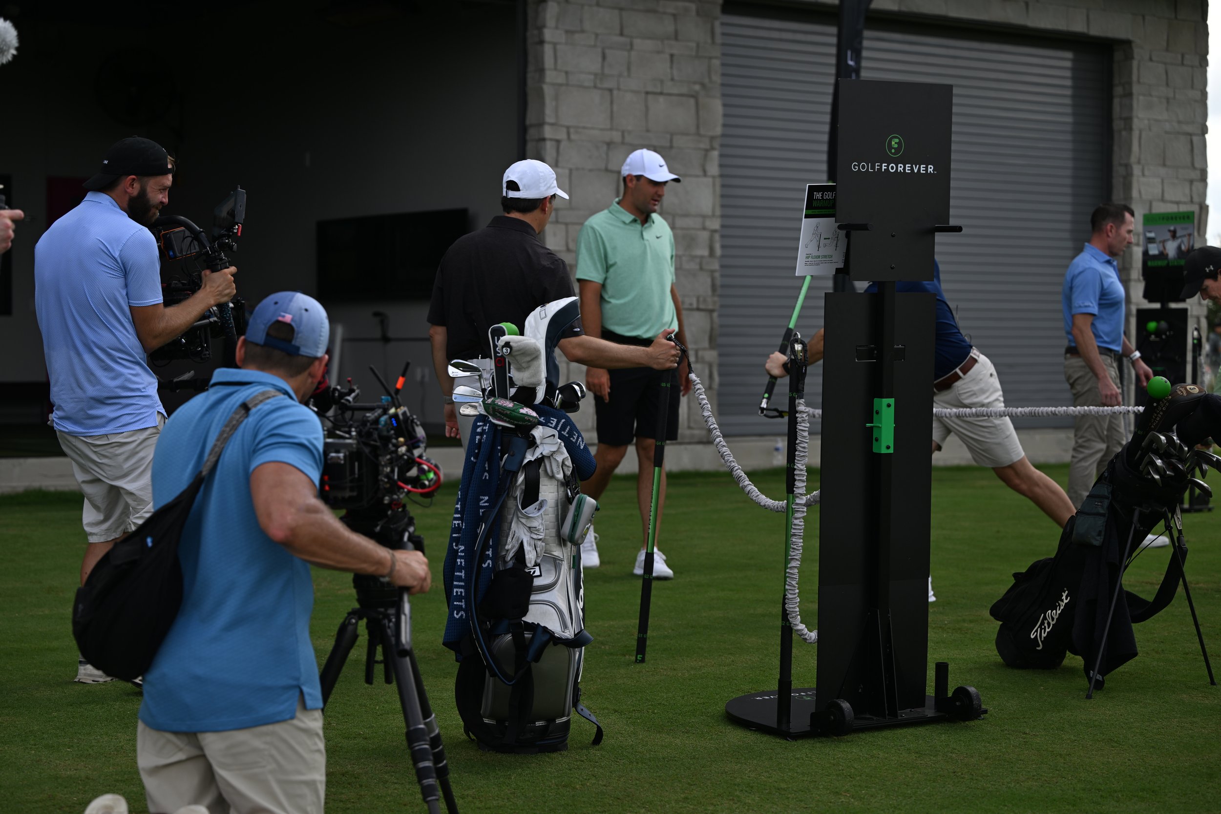 Golfers using training equipment at a driving range, surrounded by camera crew filming the activity.