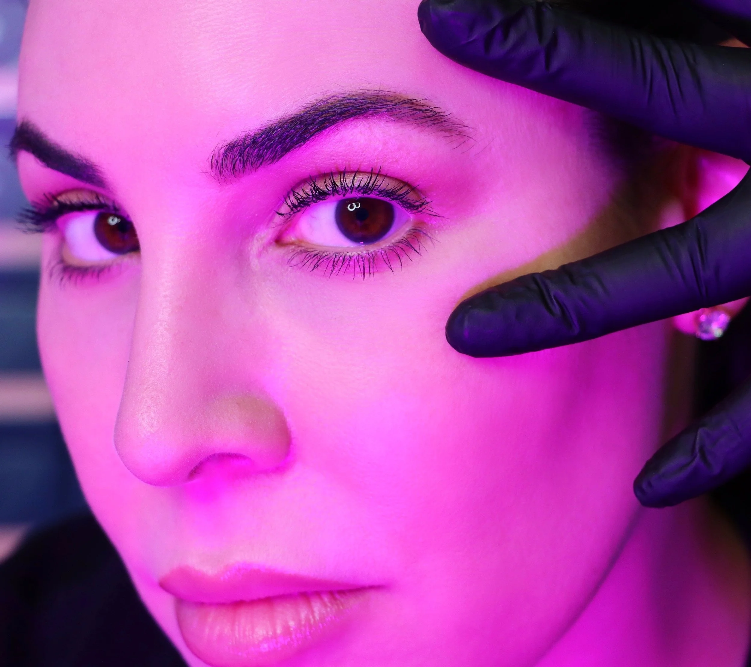 Close-up of a person with a hand wearing black gloves, touching their cheek under bright pink lighting.