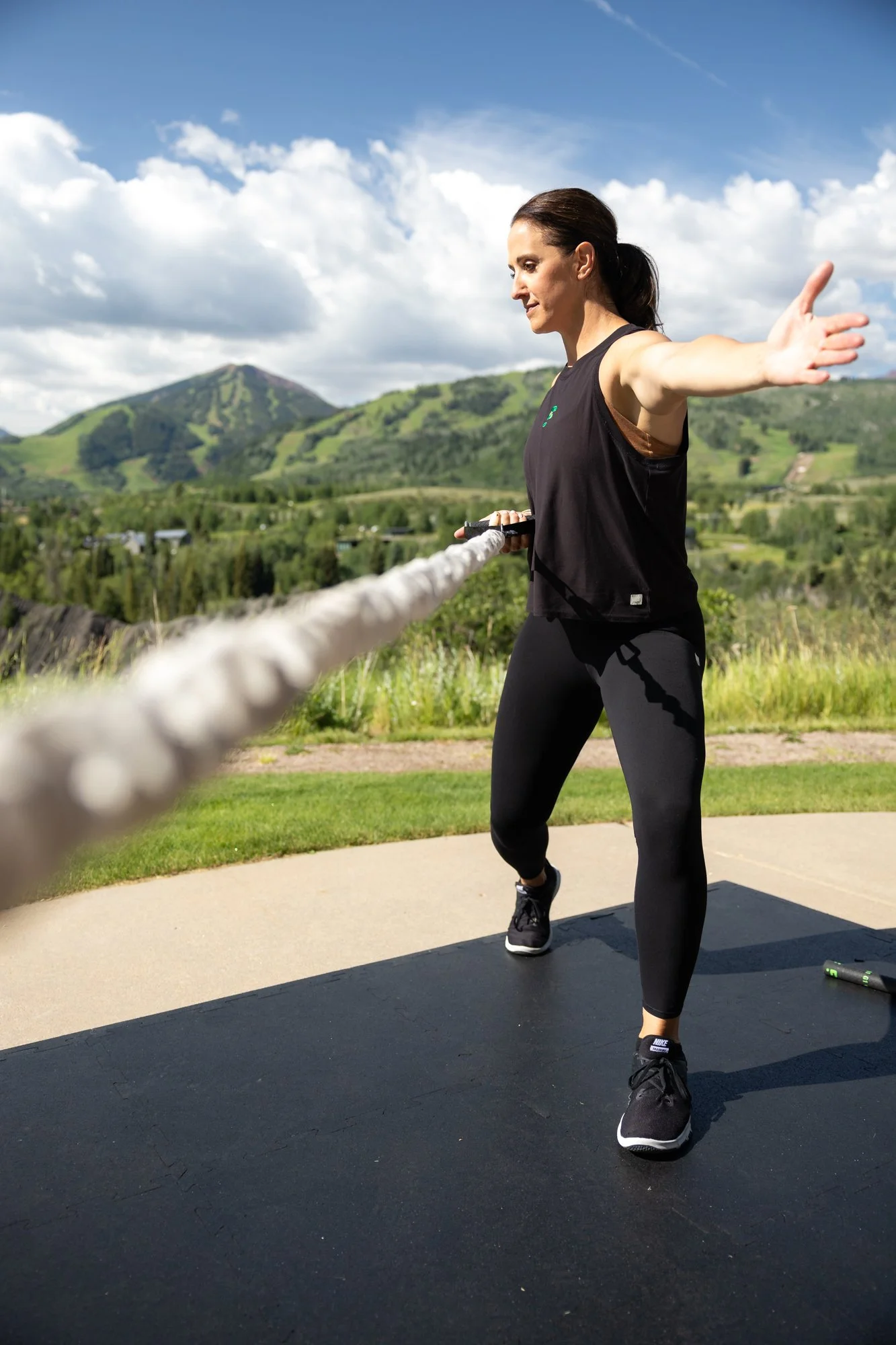 A woman exercising outdoors using battle ropes, with a scenic background of mountains and a partly cloudy sky. She is wearing a black tank top and leggings.