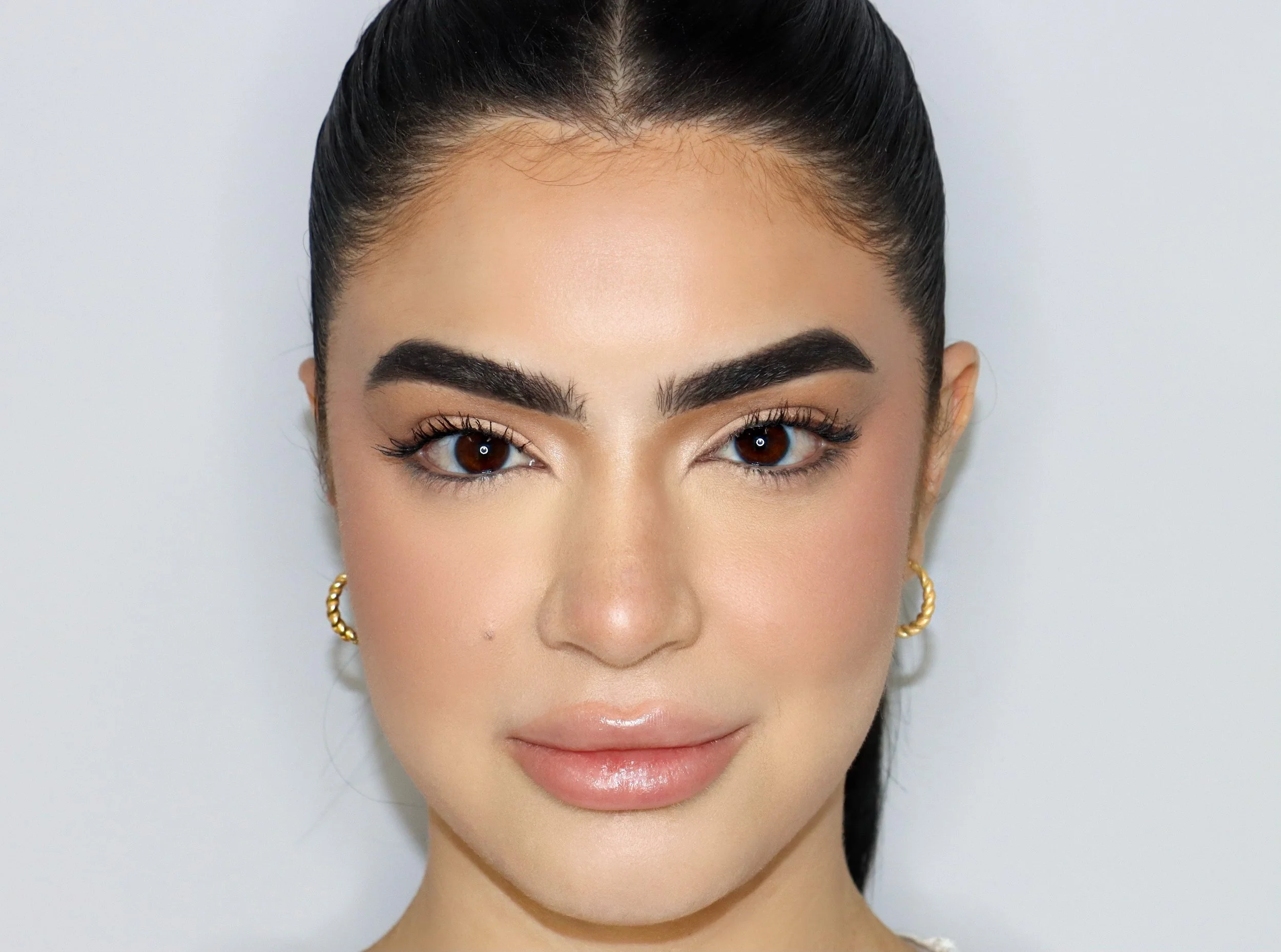 Close-up portrait of a person with dark hair, bold eyebrows, and gold hoop earrings, with a neutral background.