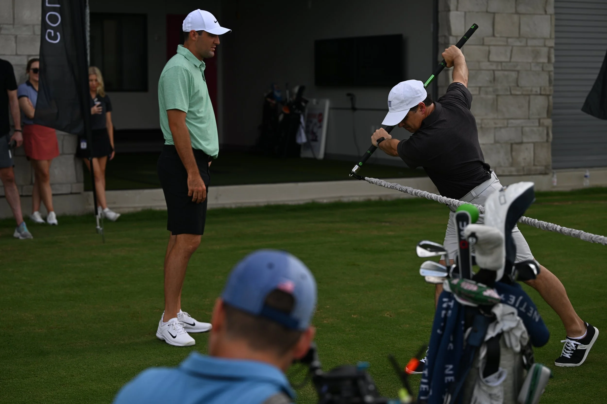 Two men on a golf course, one swinging a training device in front of the other, with spectators and golf equipment visible in the background.