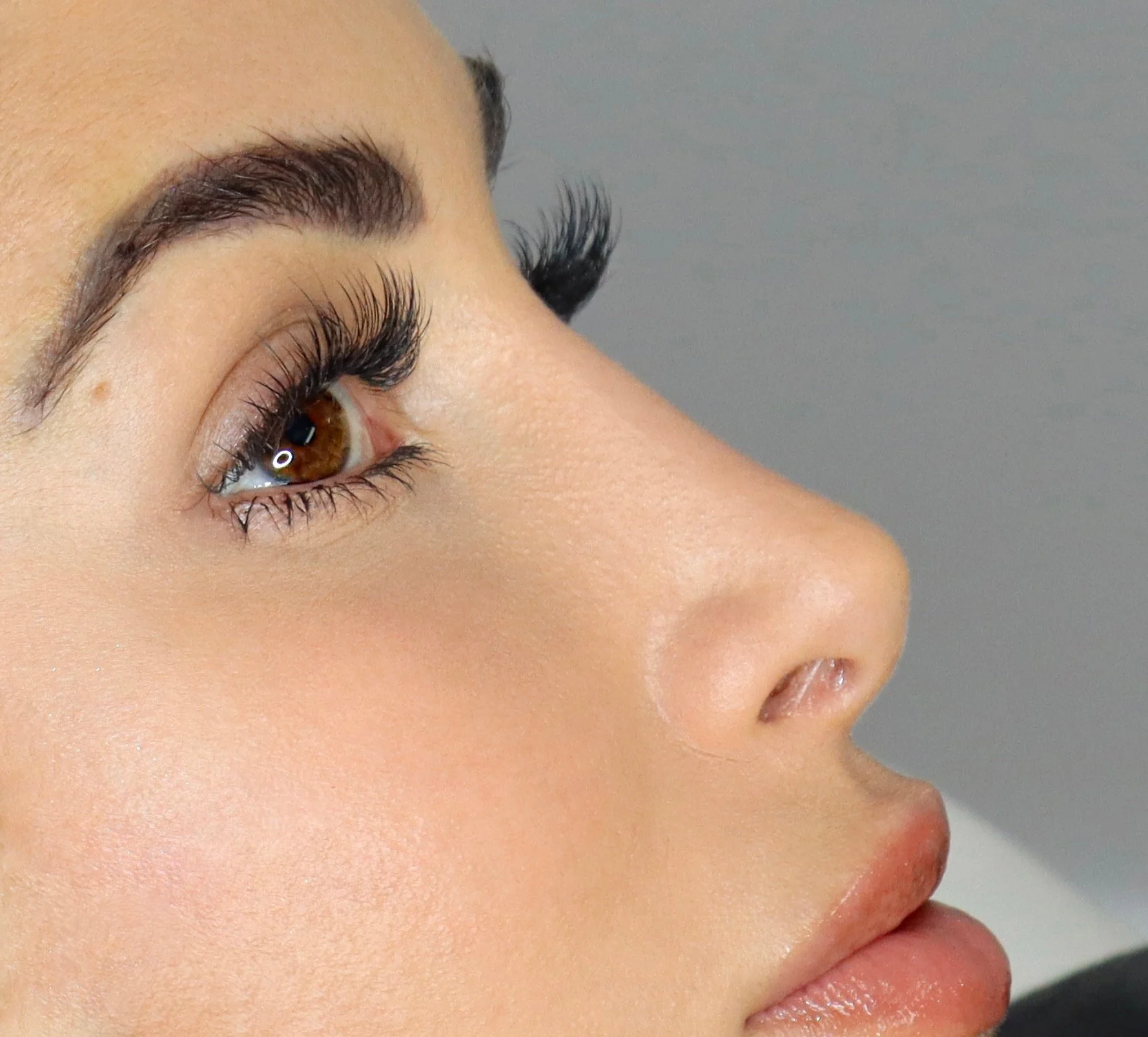 Close-up of a woman's face in profile, highlighting her eye, eyelashes, eyebrow, nose, and lips against a gray background.