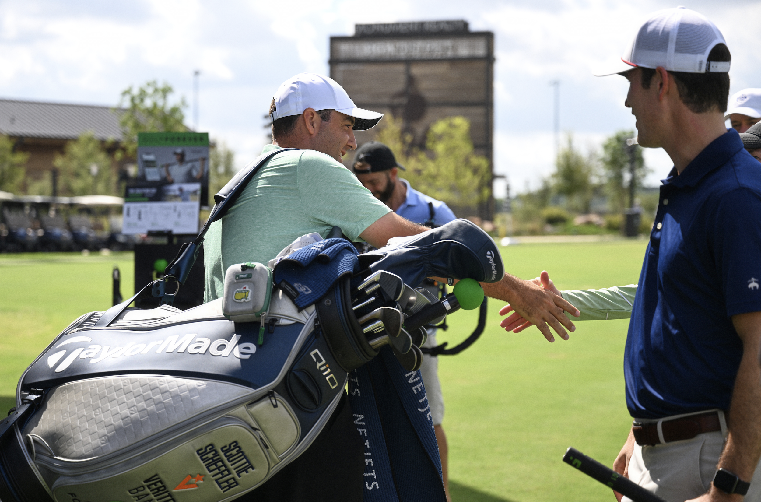 People on a golf course, one carrying a TaylorMade golf bag, shaking hands, with golf carts in the background.