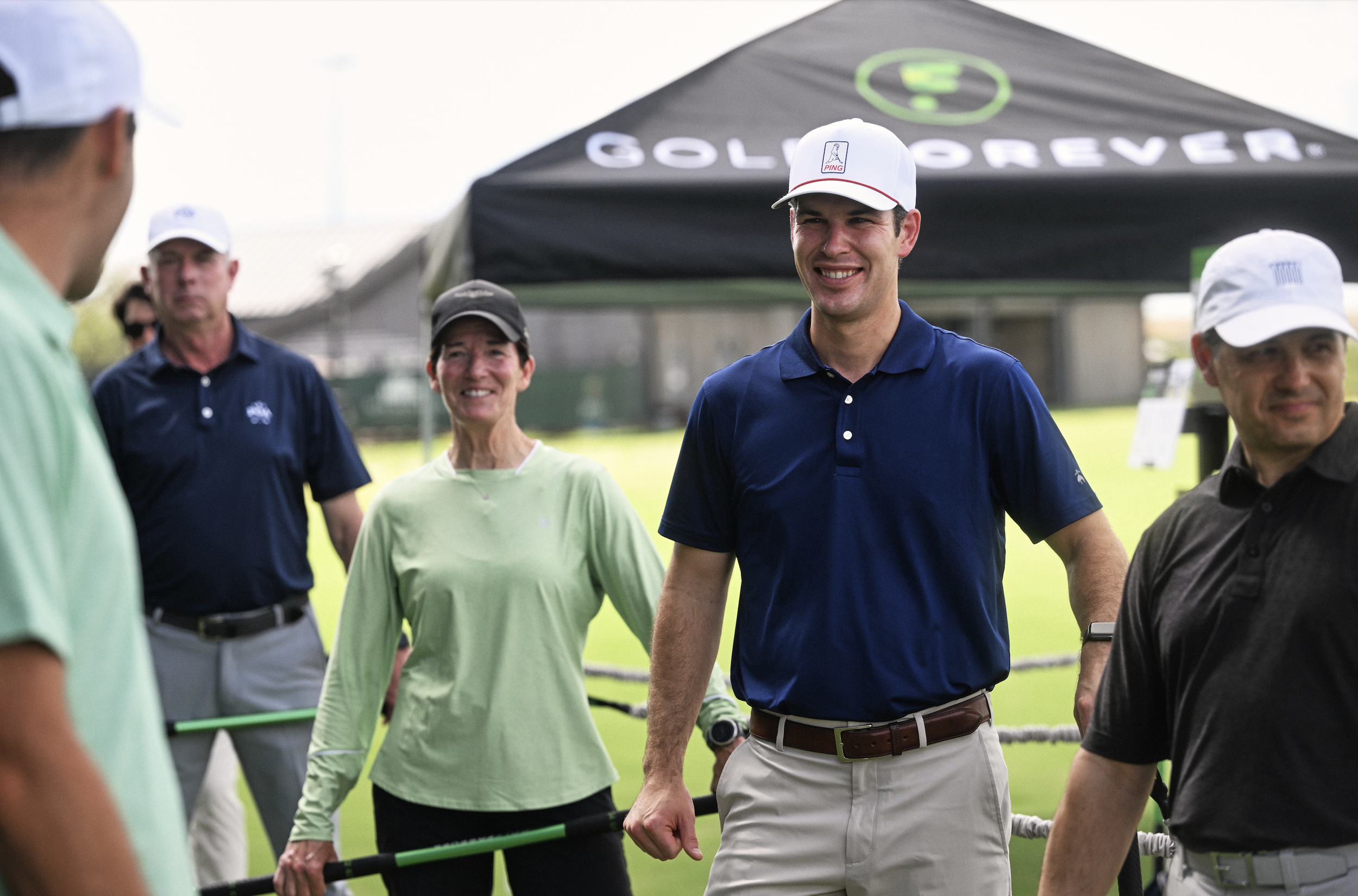 A group of people standing outdoors near a tent with "Golf Forever" logo, some wearing golf attire and caps, smiling and appearing to be enjoying a golf event or gathering.