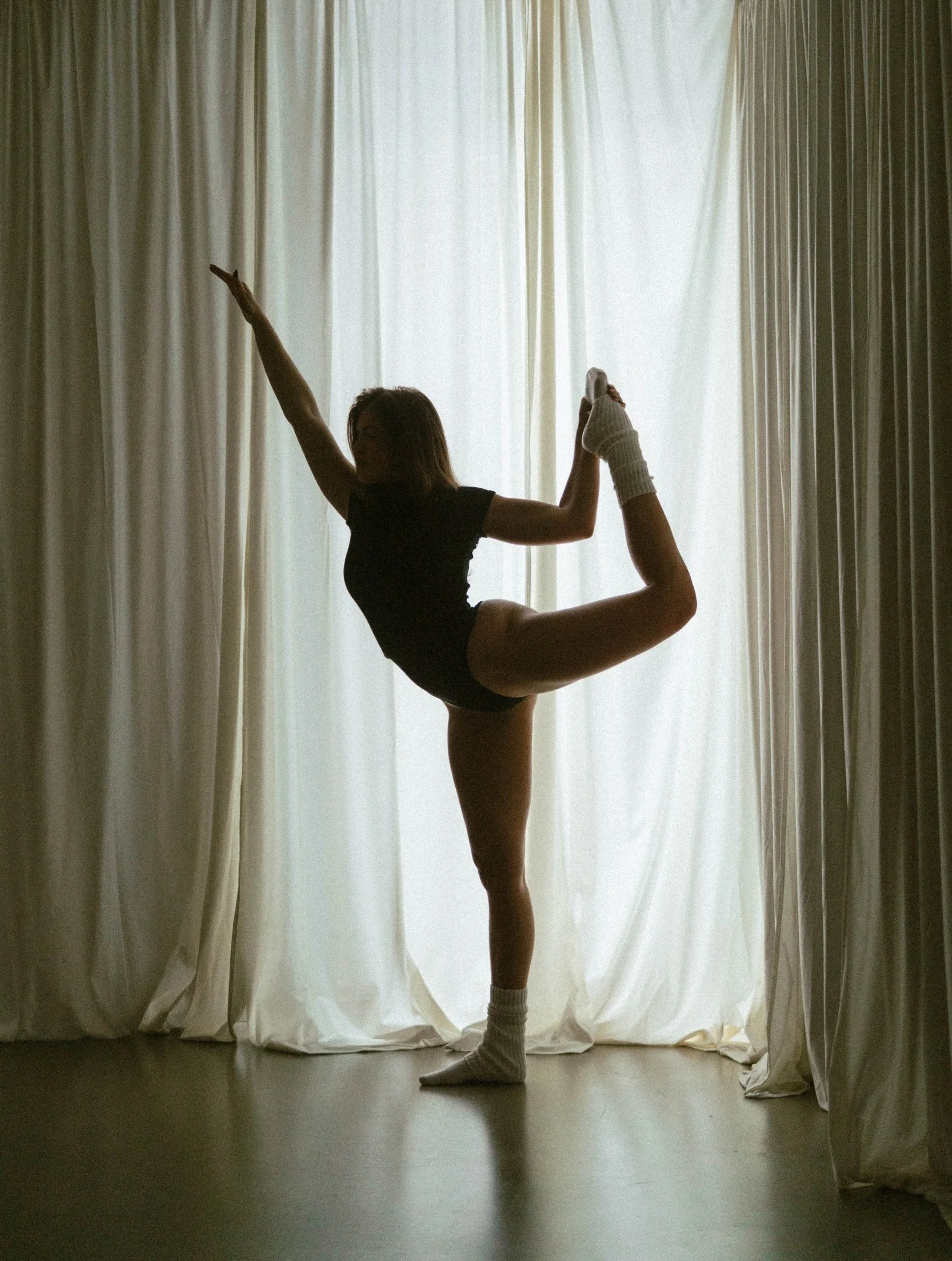 Woman in black Pilates attire balances on one leg in front of white drapery.