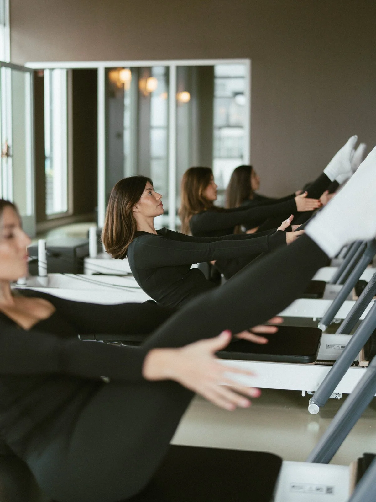 Women in black exercise clothes extend their legs during a Pilates reformer class.