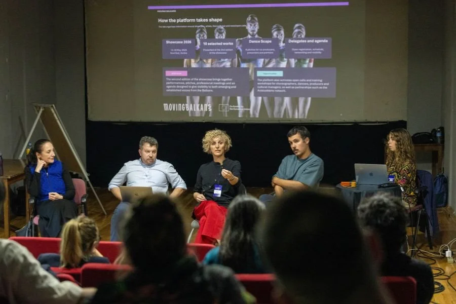 A panel of people speak in front of an audience in a spall theatre space