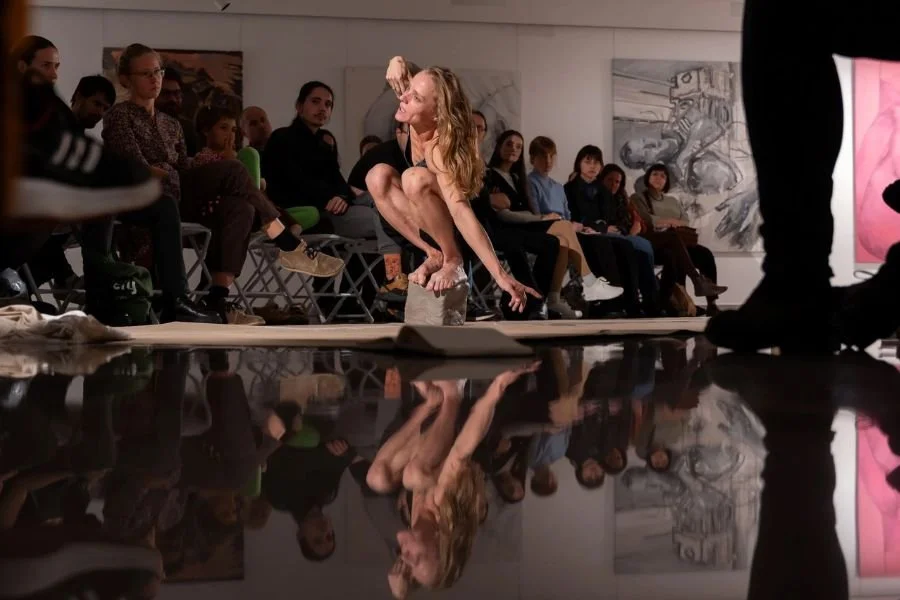 A single performer crouches and balances on a small block of concrete in a gallery space with audiences sitting and watching
