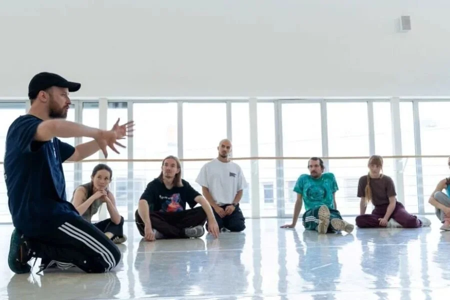 Group in a dance studio with a backdrop of windows  with light streaming in, listen to the workshop instructor