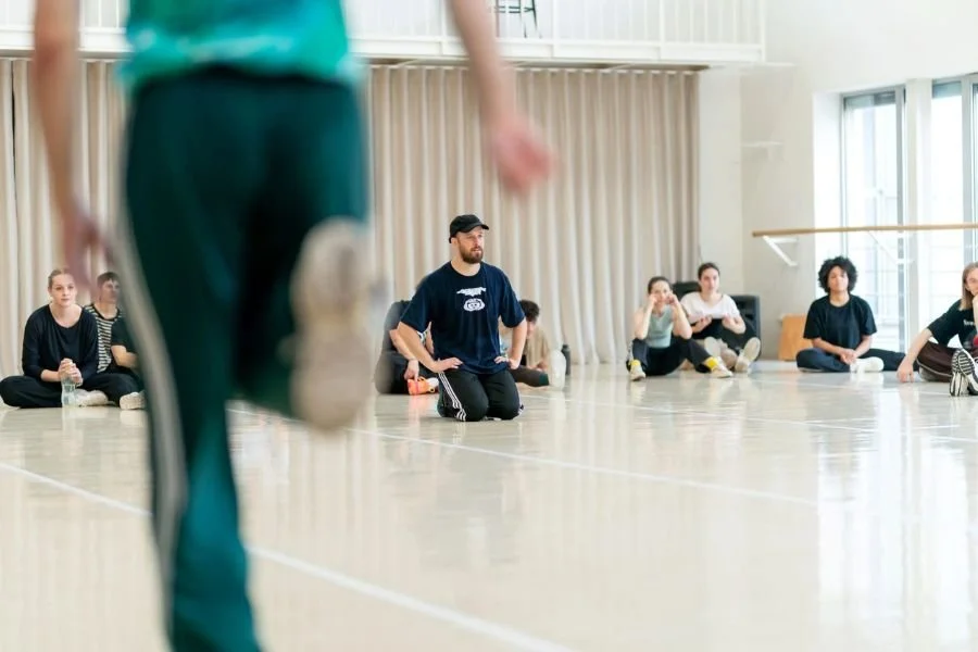Group of dance artists in a bright studio, one figure blurred in the foreground and all others further away in focus
