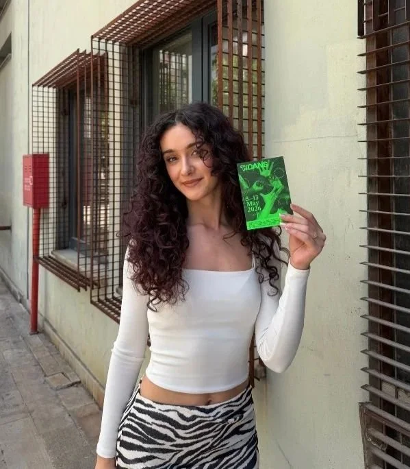 Woman with long dark curly hair, wearing a long sleeved white top and zebra print pants, holding a programme for STHLM Dans