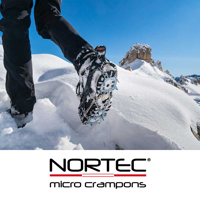 A person in black winter clothing, including gloves, is walking on snow-covered ground in a mountainous area and is holding a pair of snowshoes.