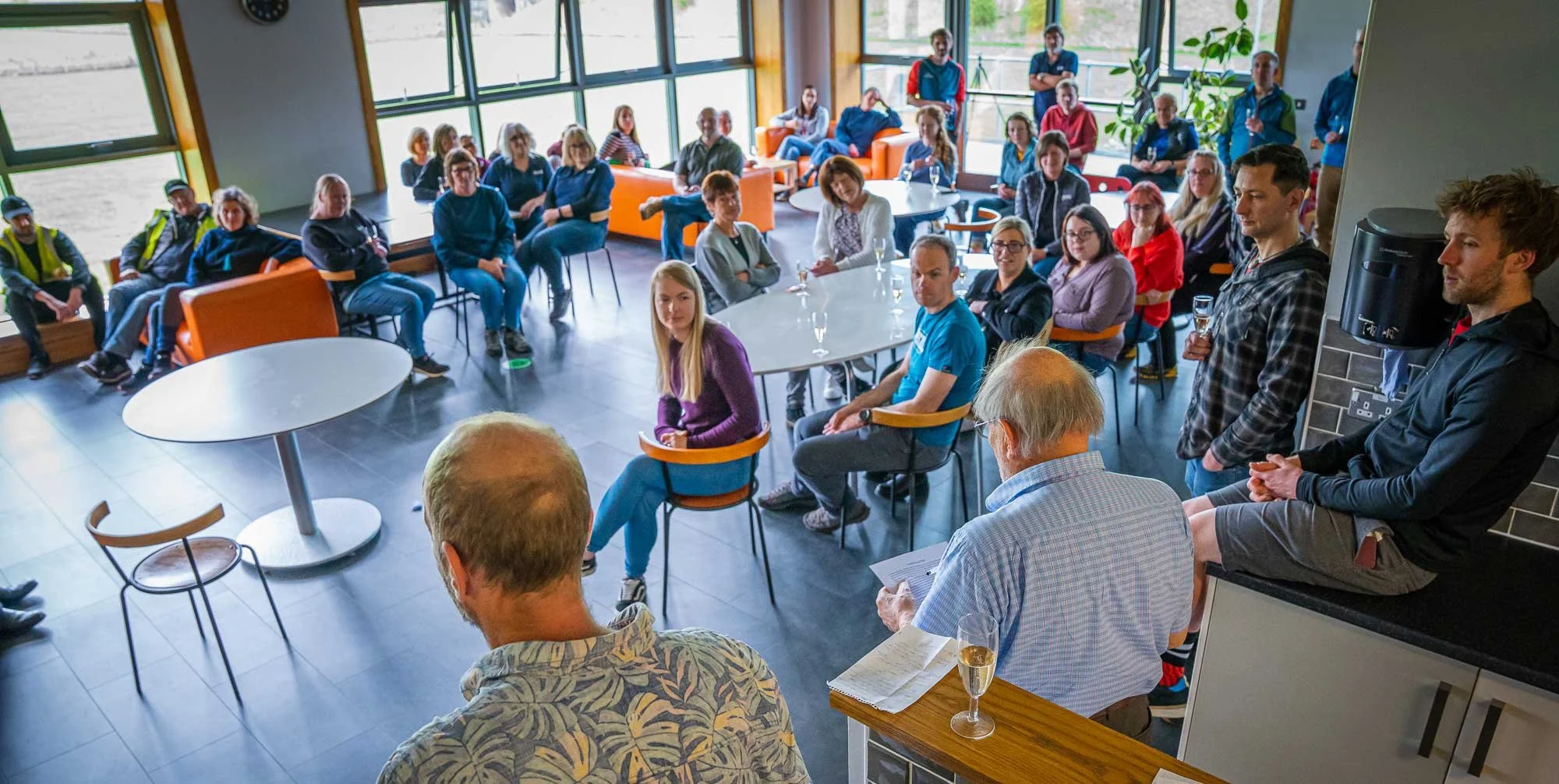 A group of people attending a meeting or presentation in a bright, modern room with large windows and orange and gray furniture. The audience is seated and listening, with some standing at the back.