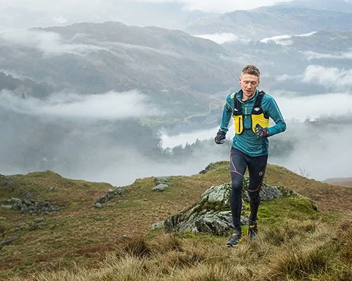 A young man trail running on a hilltop with foggy mountains in the background.