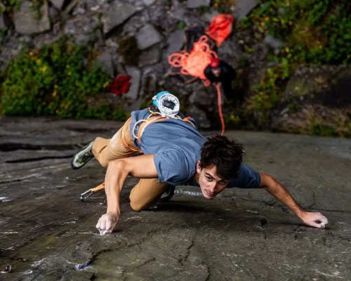 A man engaged in rock climbing on a steep, wet rock surface outdoors, with climbing gear including harness and rope.