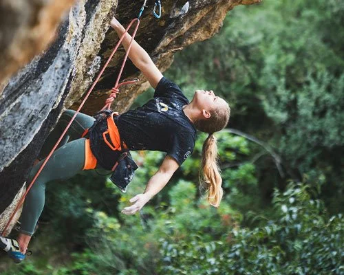 Female rock climber in black shirt and green pants reaching for holds on an outdoor climbing wall with greenery in the background.