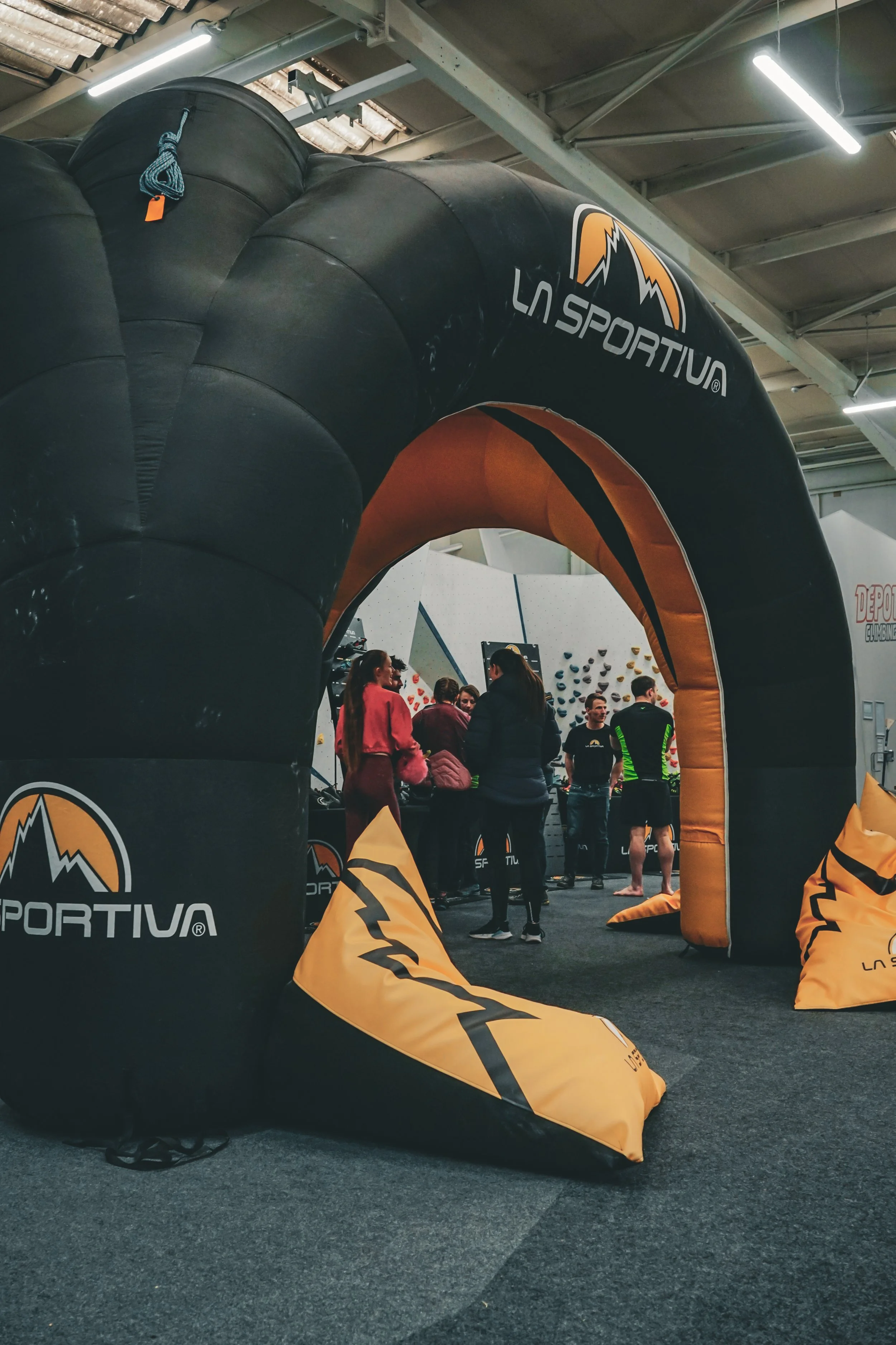 Indoor rock climbing gym with an orange and black inflatable archway branded La Sportiva at the entrance, leading to a climbing area where people are gathered and preparing for climbing activities.