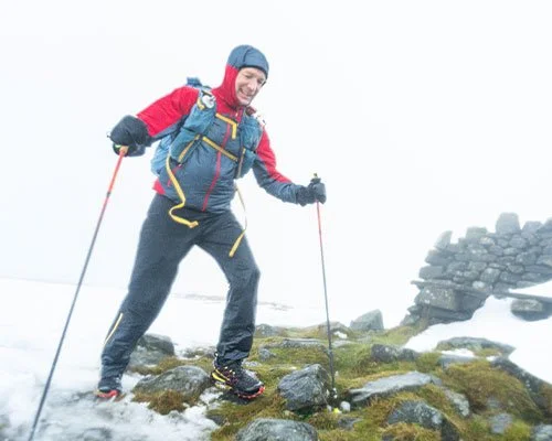 Hiker in winter gear crossing a rocky, snowy terrain with trekking poles, foggy background, and a stone cairn.