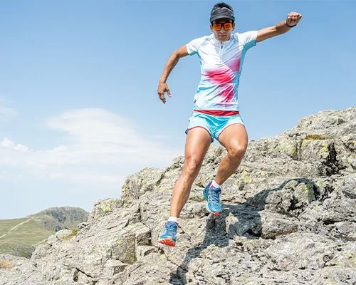 A woman in athletic wear is running or jumping over rocky terrain outdoors, with hills and a blue sky in the background.