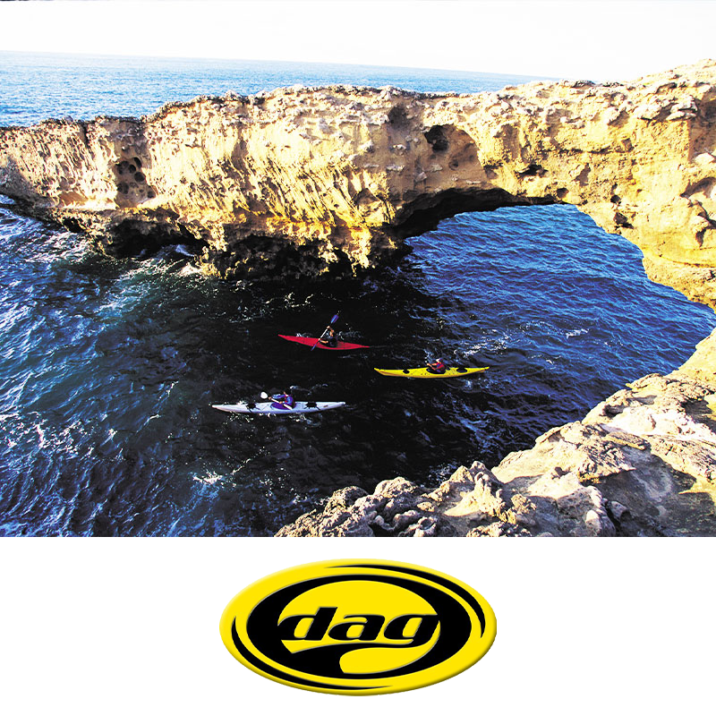 Three kayakers paddle near a natural ocean rock formation with a large arch and caves.