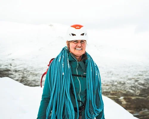 A woman wearing a white helmet and teal jacket, smiling, standing in a snowy landscape with a large sheathed climbing rope on her shoulder.