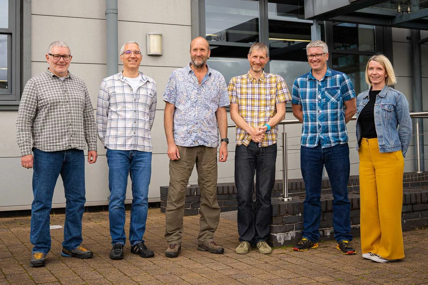 Seven people standing outside in front of a modern building, posing for a group photo. They are dressed casually and smiling.