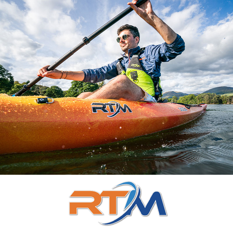 A man kayaking on a lake, wearing sunglasses and a life jacket, with green trees and hills in the background under a partly cloudy sky.