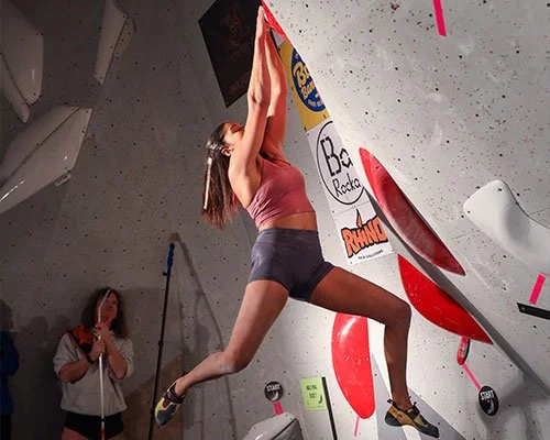 A woman in a pink sports bra and black shorts climbing an indoor rock climbing wall, with a person observing in the background.