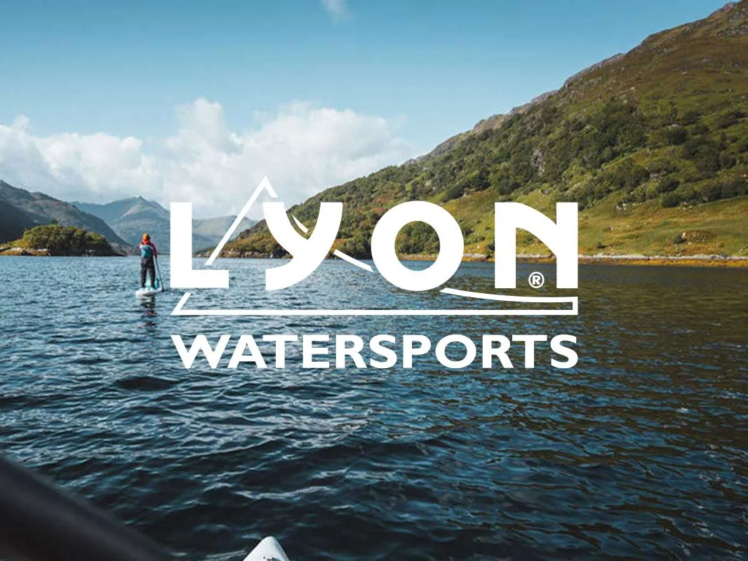 A person stand-up paddleboarding on a calm lake surrounded by green mountains under a blue sky with clouds, with the logo 'Lyons Watersports' overlaid.