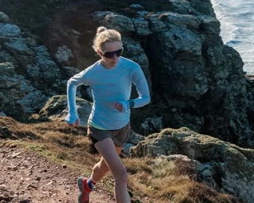 A woman running along a rocky coastal trail, wearing sunglasses, a light blue long sleeve shirt, shorts, and running shoes.