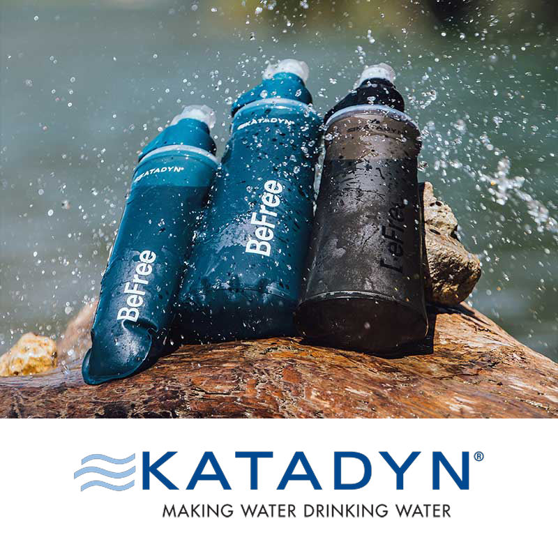 Three Katadyn Befree water bottles, two blue and one black, placed on a log near water with splashes, with a Katadyn logo and slogan below.