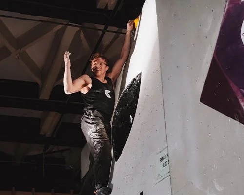 A person in athletic clothing climbing an indoor rock wall with a smile and raised arm for celebration.
