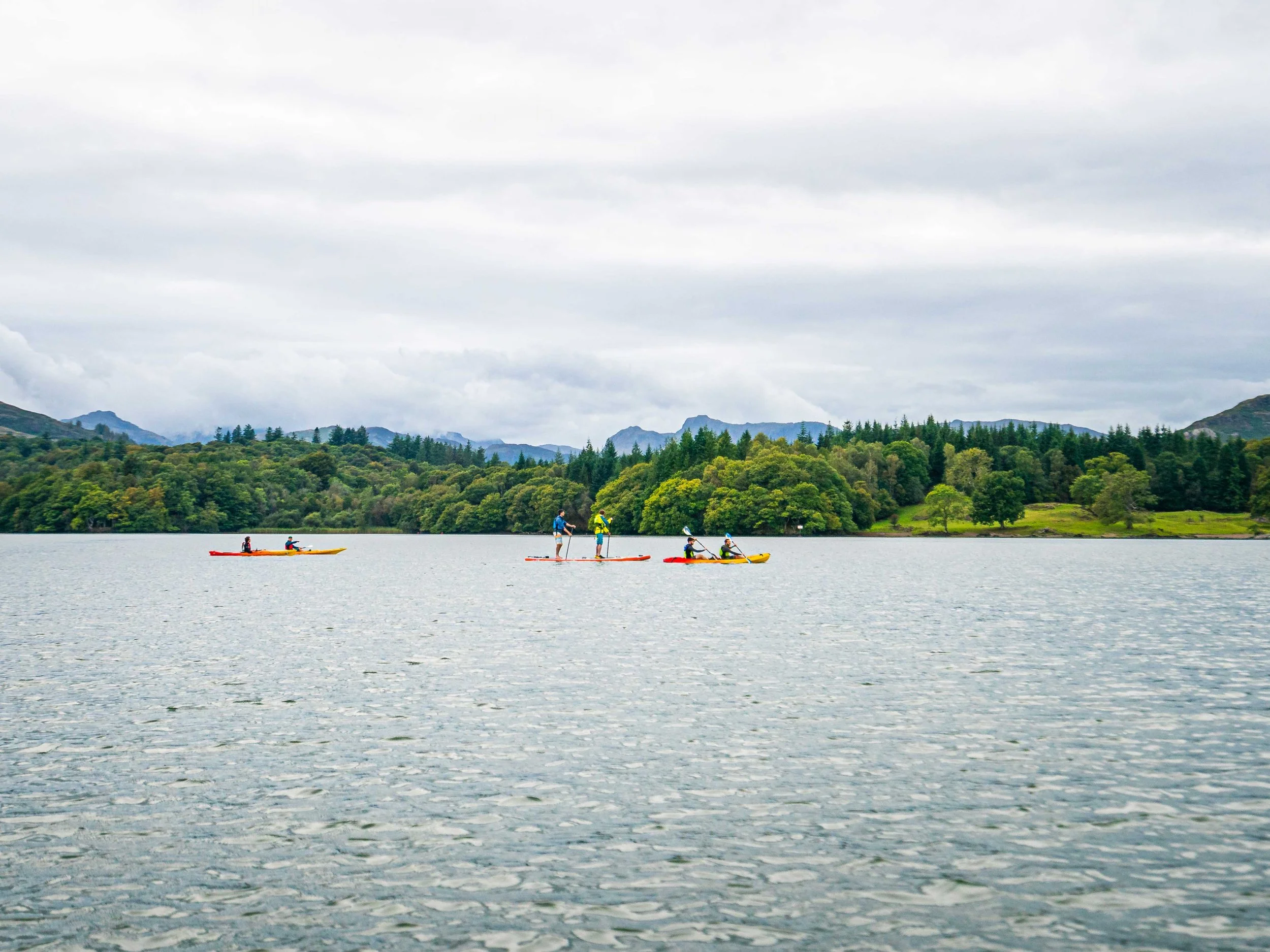 People paddleboarding and kayaking on a lake with a forested shoreline and mountains in the background under a cloudy sky.