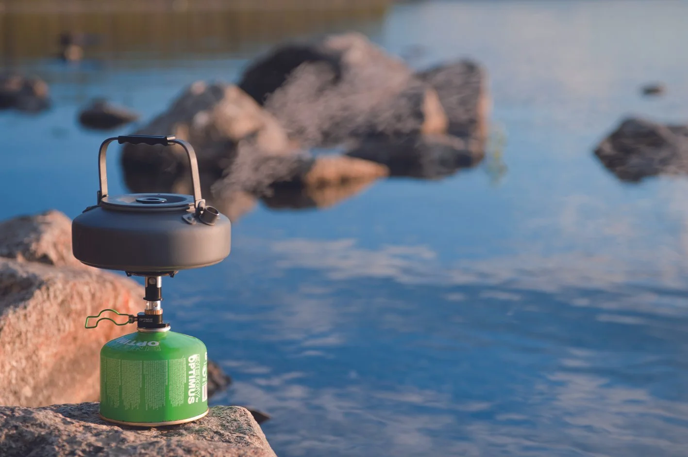 Portable camping stove with a green fuel canister placed on a rock near a body of water with large rocks in the background.