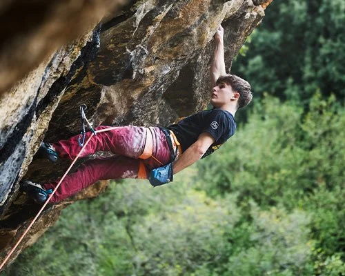 Young man rock climbing outdoors on a steep, overhanging rock face, with trees in the background.