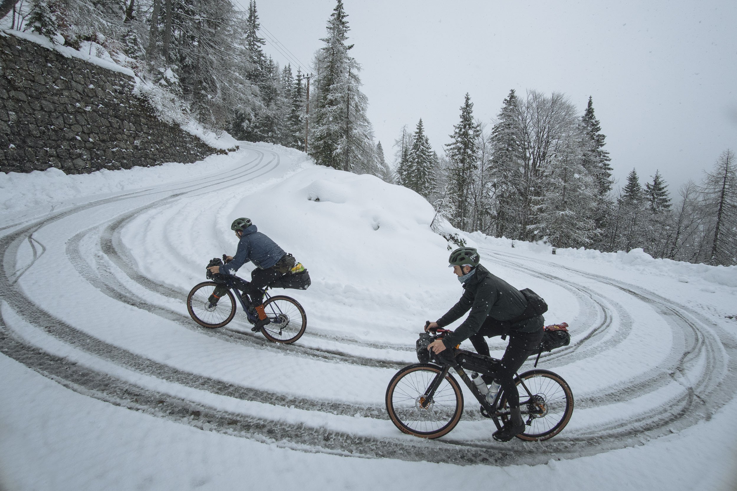 Two cyclists riding mountain bikes on a snow-covered winding road with snowy trees in the background.