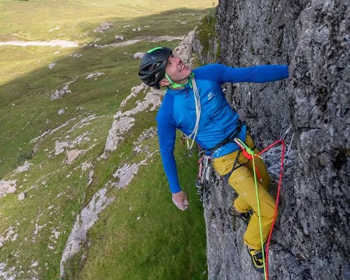 Climber ascending a steep rock face in a green mountainous landscape, wearing a helmet, blue jacket, and yellow pants.