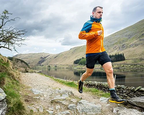 Man running on a rocky trail beside a lake in hilly countryside.
