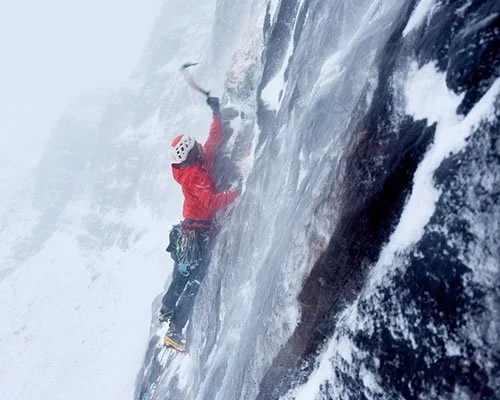 Climber in red jacket and helmet ascending an icy, snow-covered rock face with a hammer in hand.
