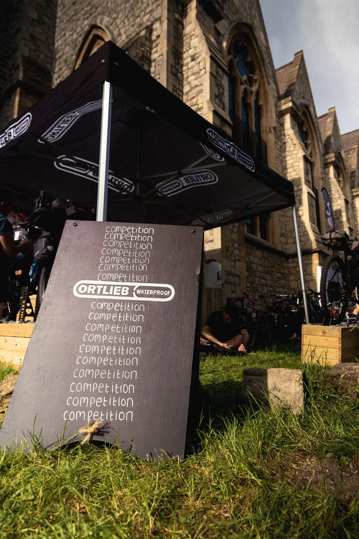 A chalkboard sign with the word 'competition' written multiple times and the 'Ortlieb Waterproof' logo, placed outdoors in front of a historic stone building with large windows, under a black canopy, with bicycles and people in the background.