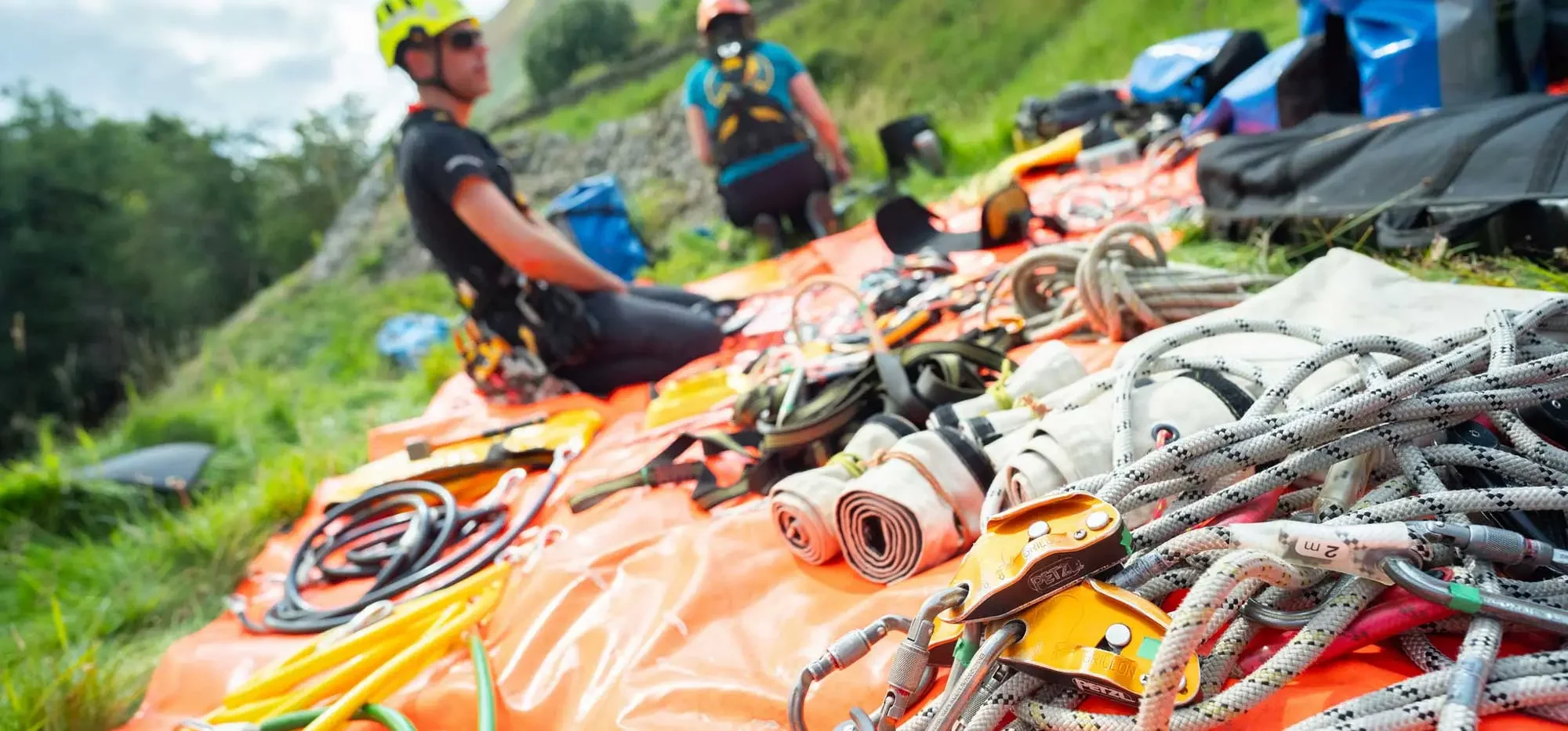 Climbing gear laid out on an orange tarp outdoors, with two climbers in helmets and harnesses in the background.