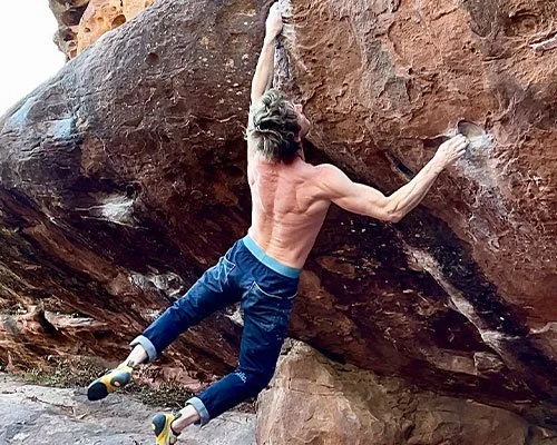 Man with long hair and no shirt rock climbing on a large brown boulder outdoors.